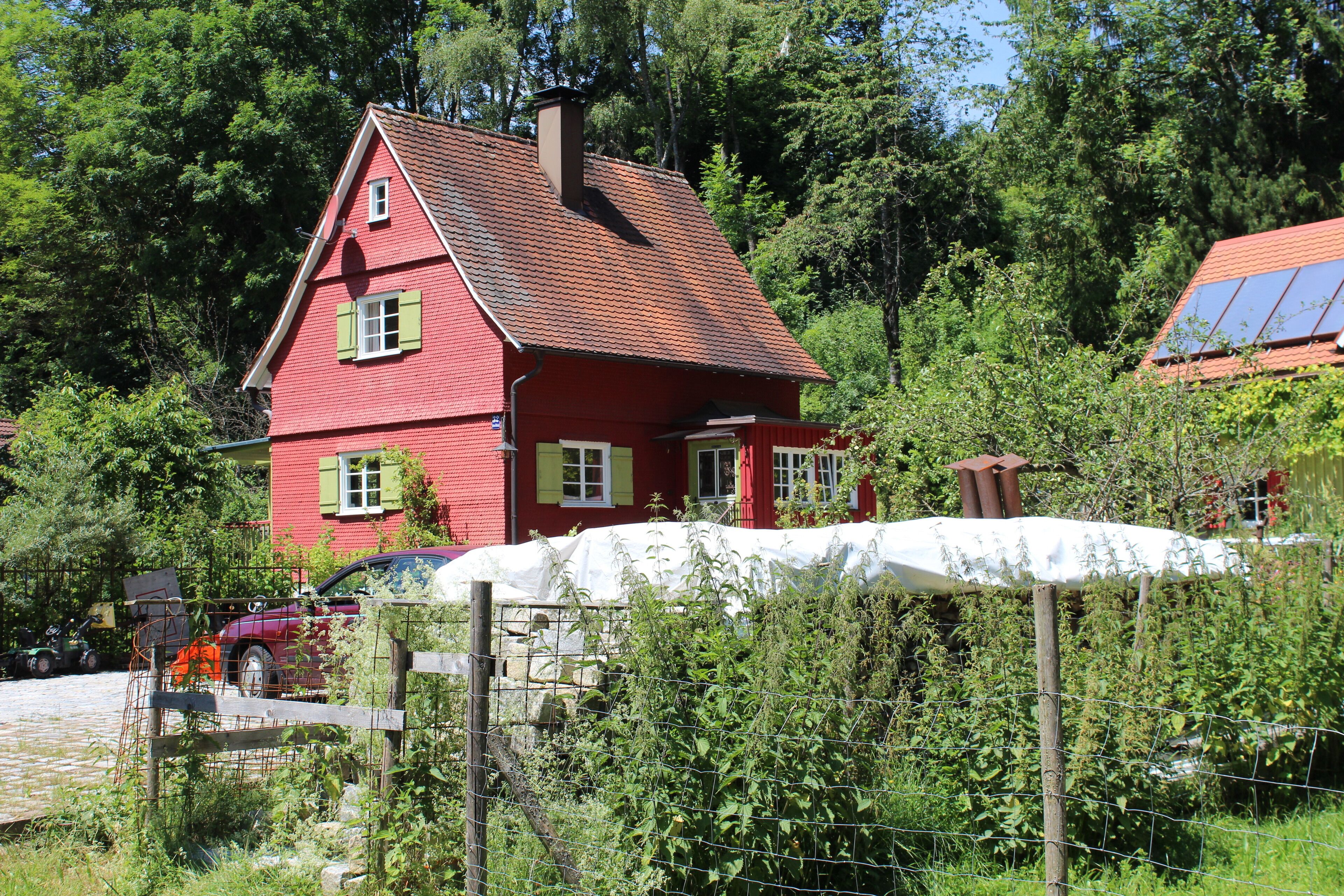 A red house with a sloped roof, surrounded by greenery and a fence.