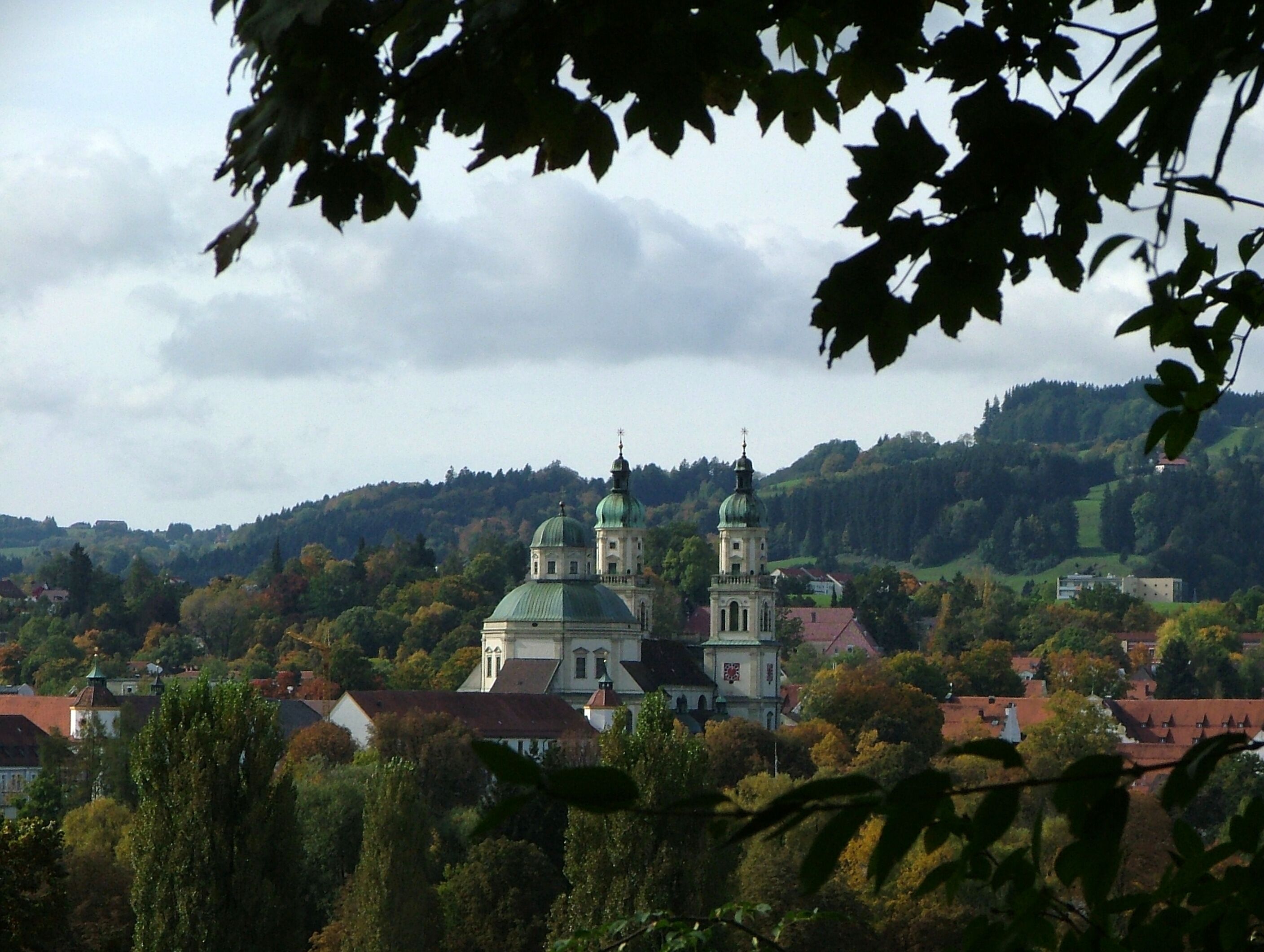 A town with two prominent green-roofed domed buildings, surrounded by autumn-colored trees and hills in the background.