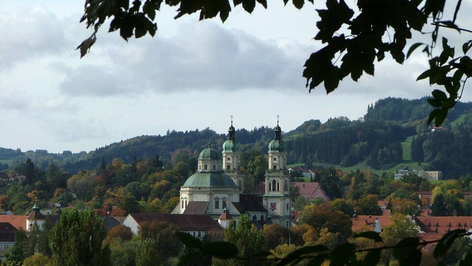 A town with two prominent green-roofed domed buildings, surrounded by autumn-colored trees and hills in the background.