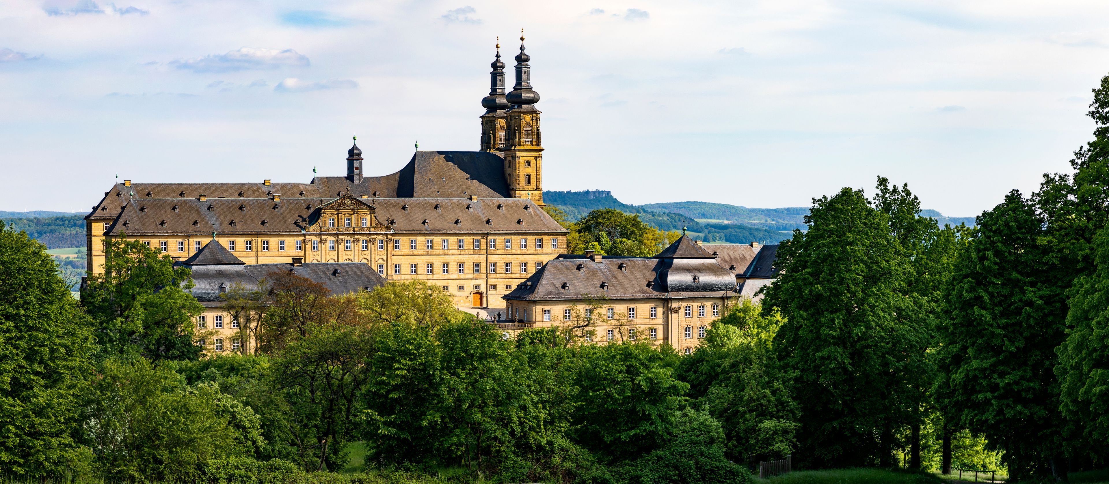 A large historic building with multiple towers and a prominent clock tower, surrounded by lush green trees and hills in the background.