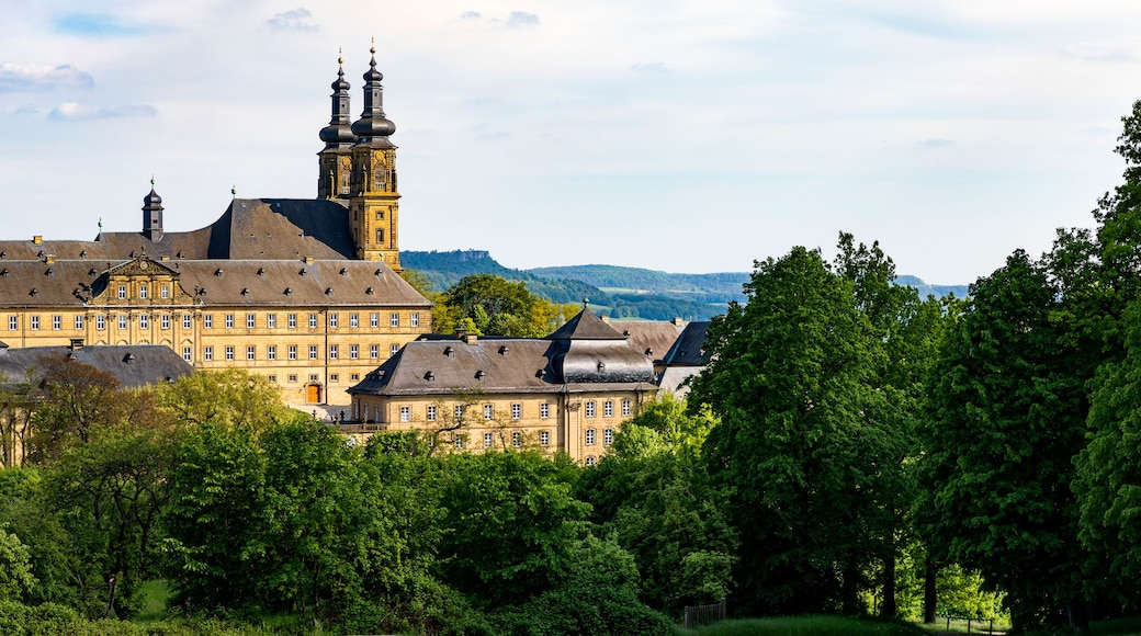 A large historic building with multiple towers and a prominent clock tower, surrounded by lush green trees and hills in the background.