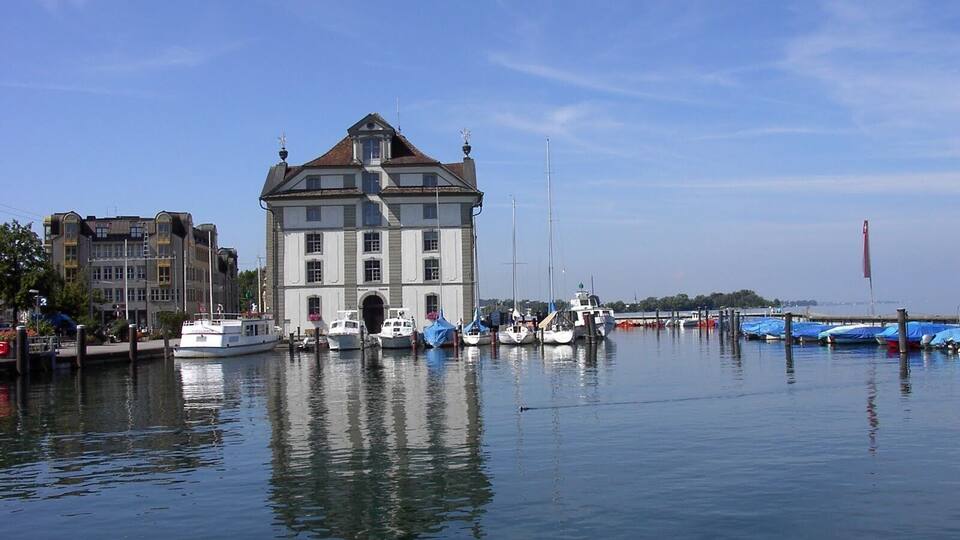 A historic building with a large arched entrance, surrounded by calm water with several boats docked.