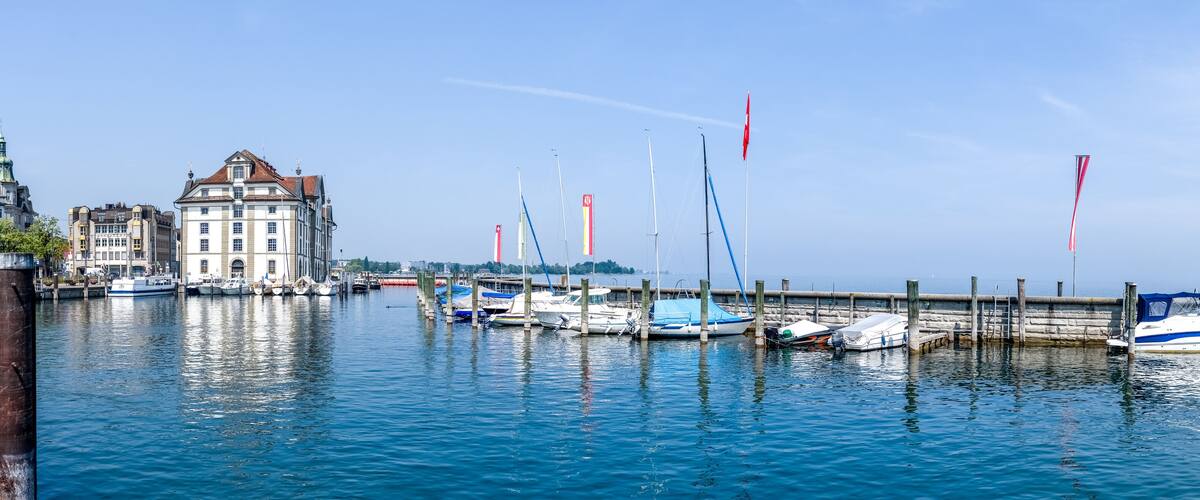 A marina with boats docked along a waterfront, a stone pier, and historic buildings in the background.