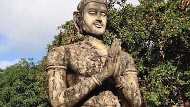 A statue of a deity with hands in a prayer position, set against a backdrop of trees and a clear sky.