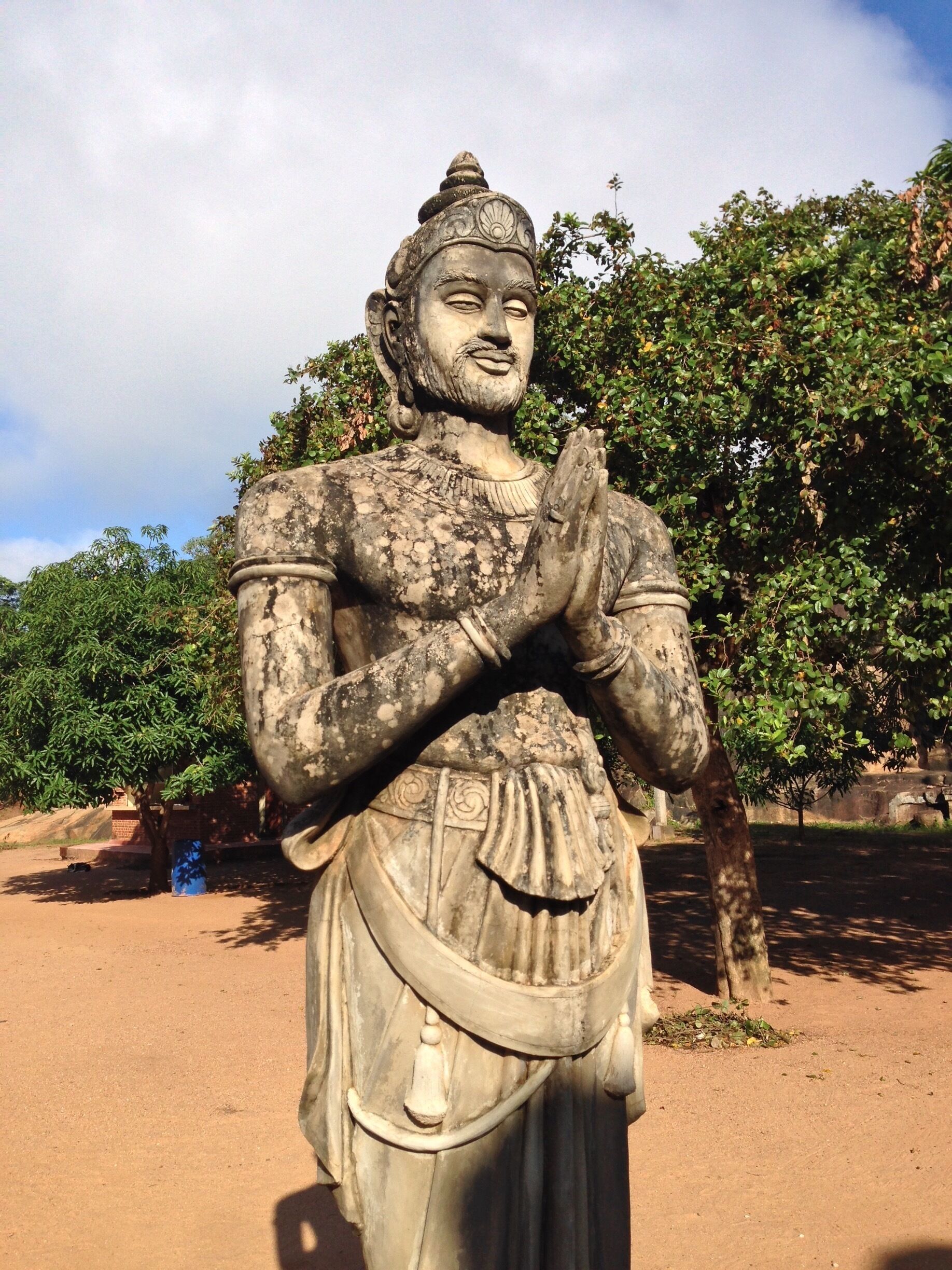A statue of a deity with hands in a prayer position, set against a backdrop of trees and a clear sky.