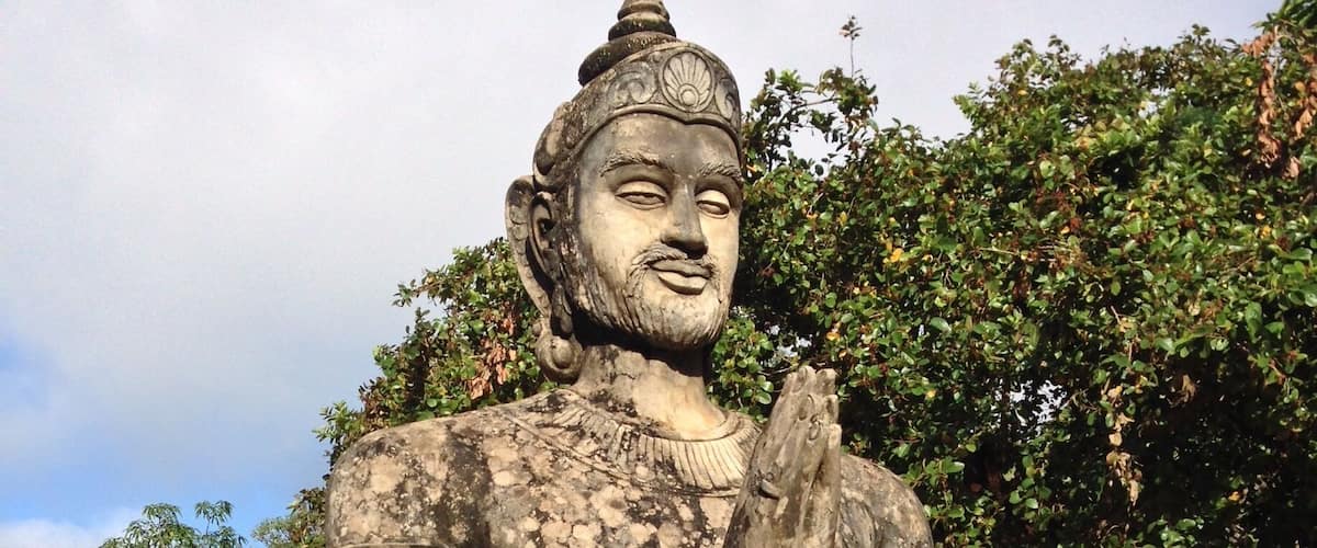 A statue of a deity with hands in a prayer position, set against a backdrop of trees and a clear sky.