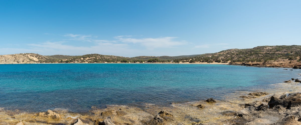 A coastal landscape with clear blue waters, rocky shorelines, and hilly terrain with sparse vegetation.