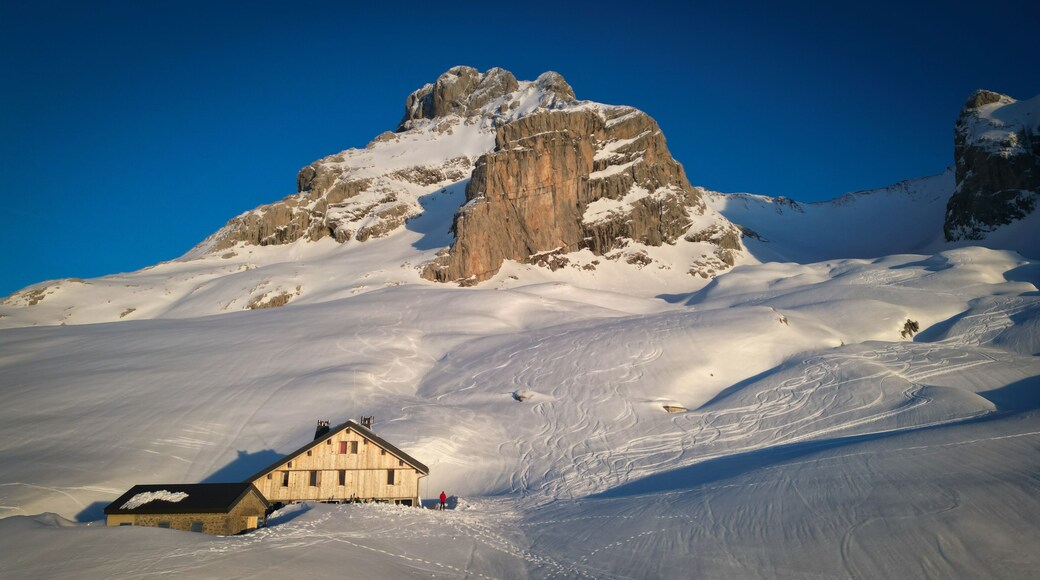 Aerial view of a Refuge de la Pointe Percee nestled in the crisp, snow-laden valley beneath the towering, rocky Pointe Percee, Le Grand-Bornand, Auvergne-Rhone-Alpes, France.