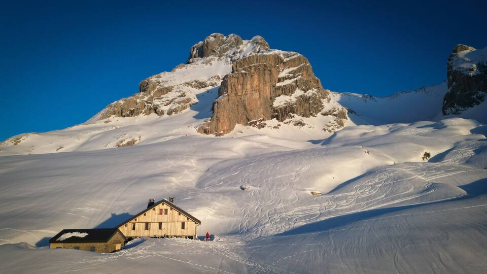 A snow-covered mountain with a prominent rocky peak, a wooden chalet, and ski tracks.