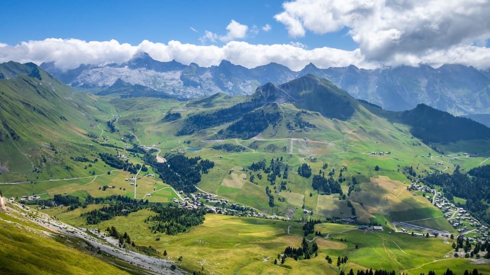 A mountainous landscape with a village nestled in a valley, surrounded by lush greenery and distant peaks.