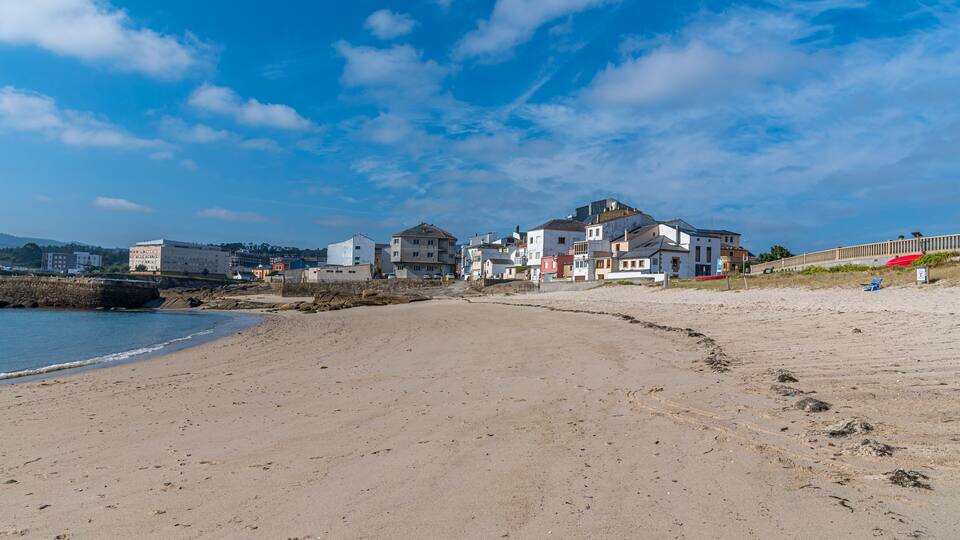 A sandy beach with footprints, a stone wall, and buildings in the background.