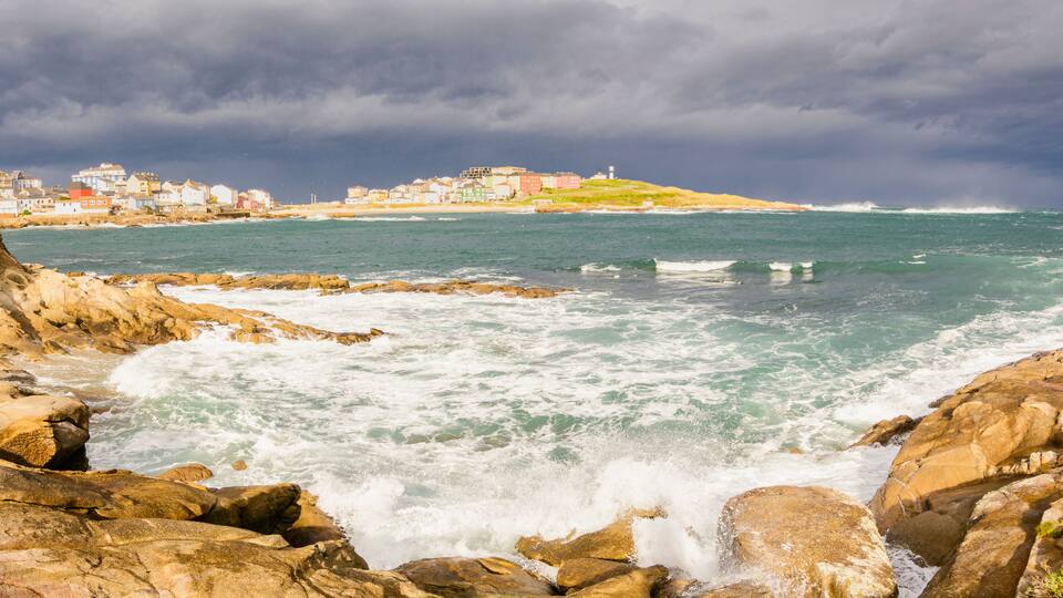 A rocky coastline with waves crashing against the rocks, a lighthouse on a hill, and a town with colorful buildings in the background.