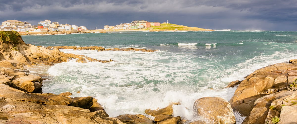 A rocky coastline with waves crashing against the rocks, a lighthouse on a hill, and a town with colorful buildings in the background.