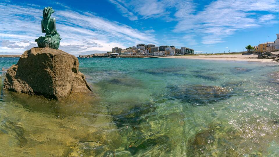 A clear coastal scene with a statue on a rock, a sandy beach, and buildings in the background.