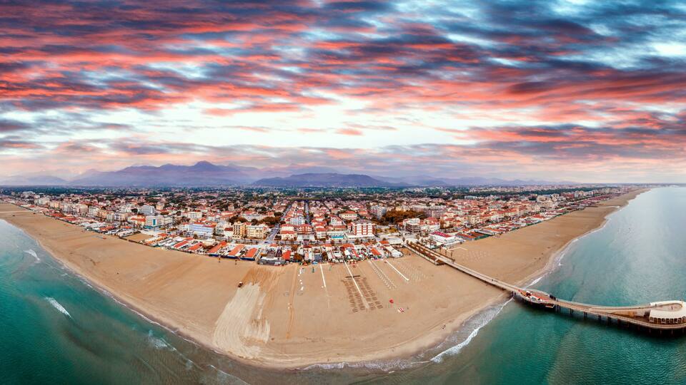 A coastal cityscape with a beach, a pier, and mountains in the distance.
