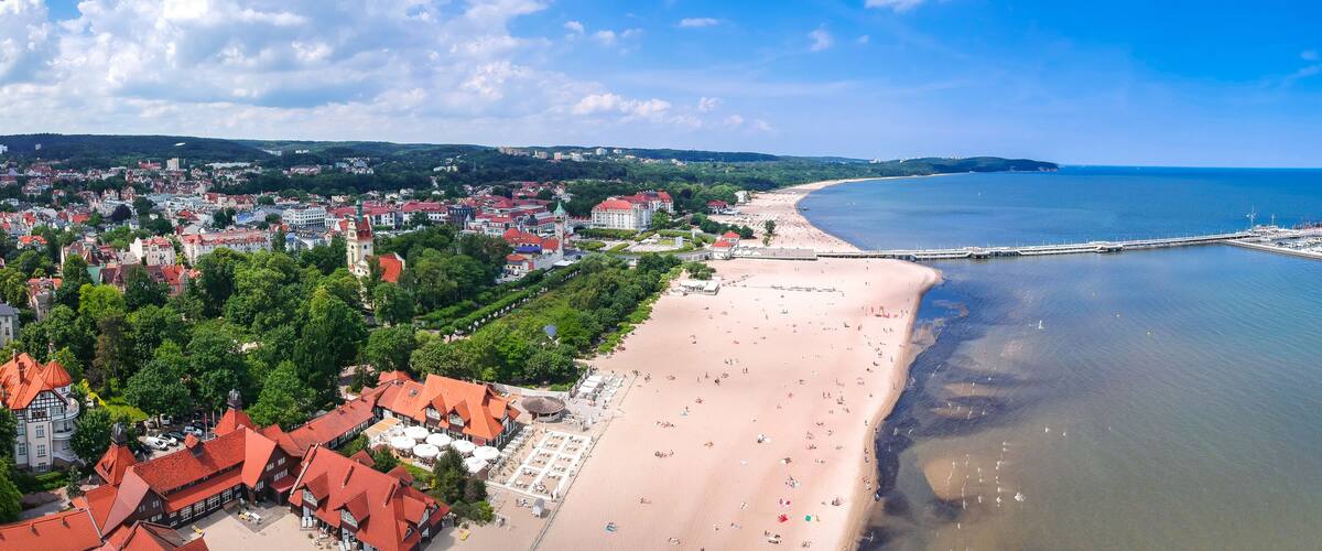 A coastal cityscape with a sandy beach, a pier, and red-roofed buildings.