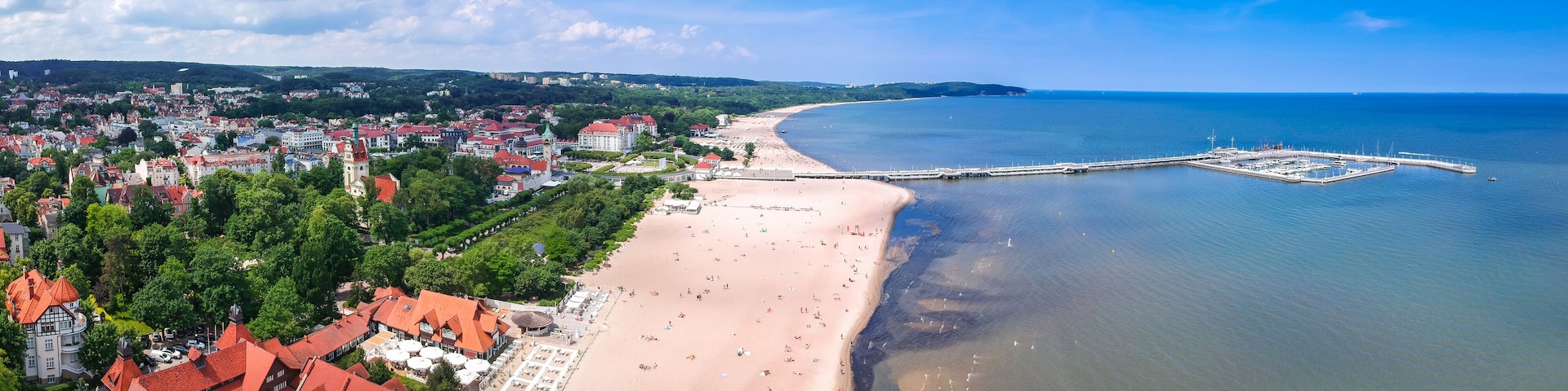 A coastal cityscape with a sandy beach, a pier, and red-roofed buildings.
