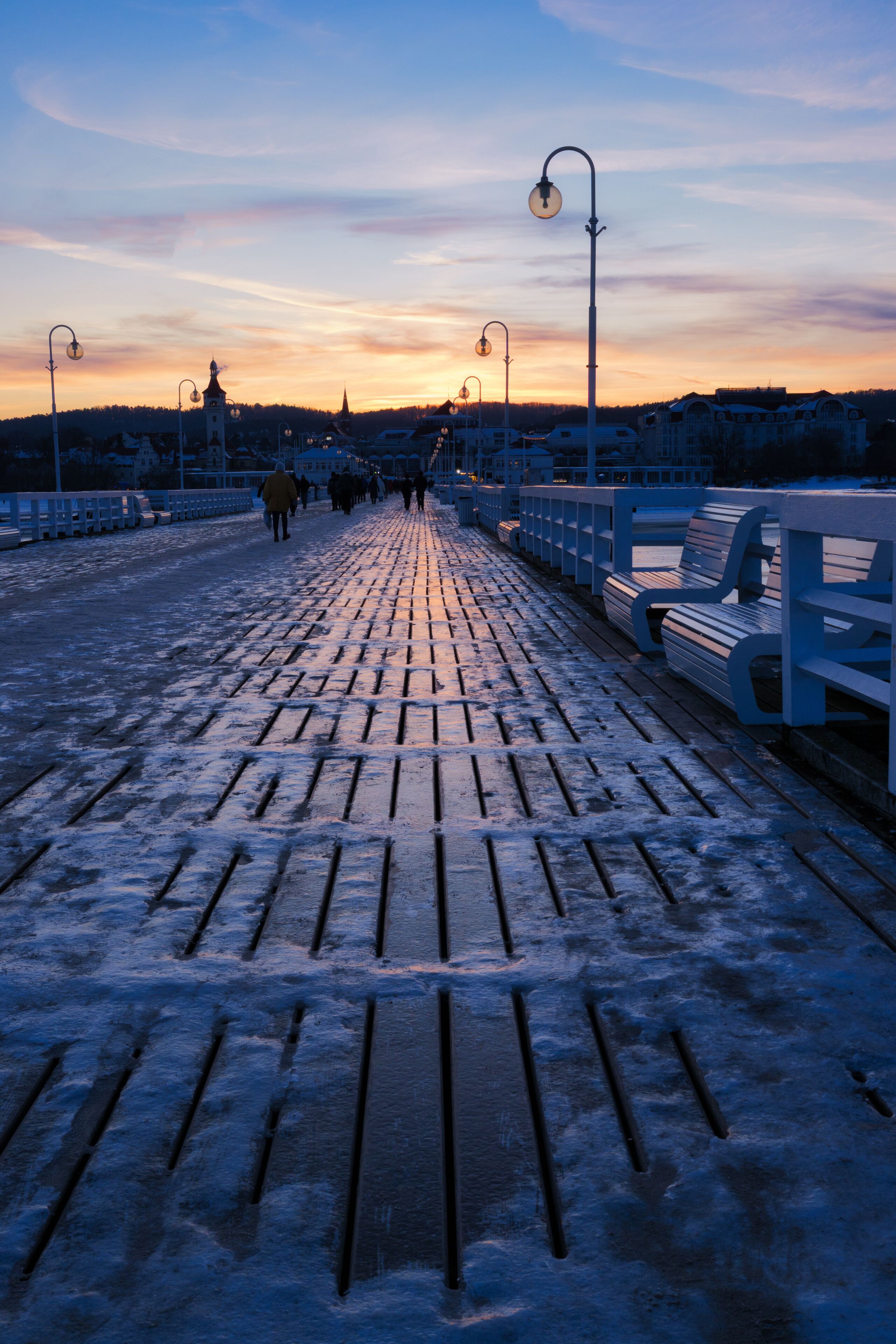 A snow-covered boardwalk with benches and street lamps, leading towards a distant town with a church spire.