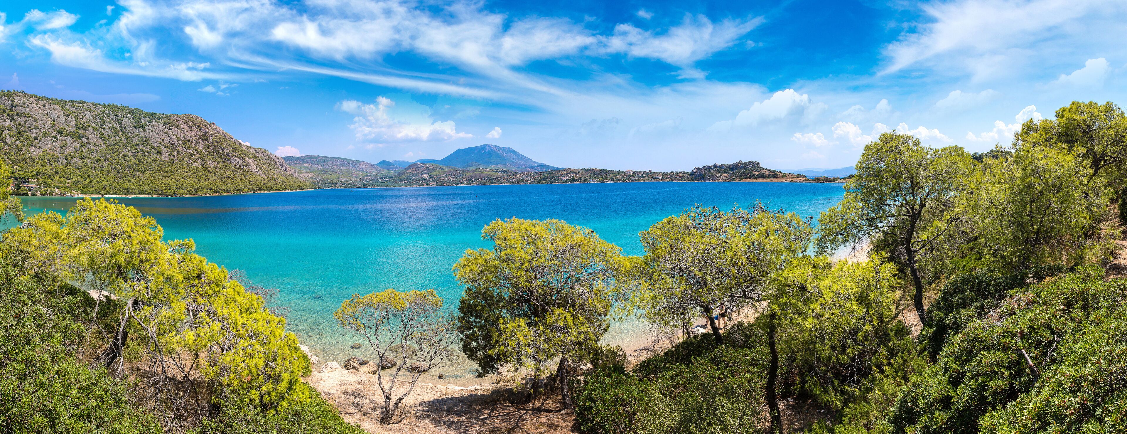 A coastal landscape with clear turquoise waters, green hills, and dense pine trees.
