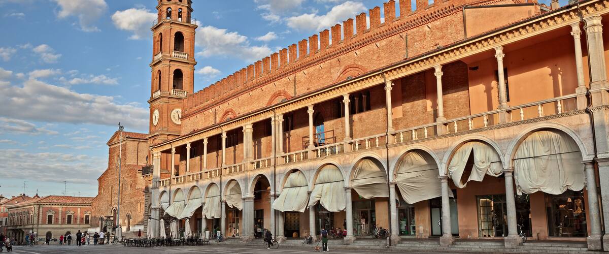 A historic square with a prominent clock tower and a building with arched windows and a tiled roof.