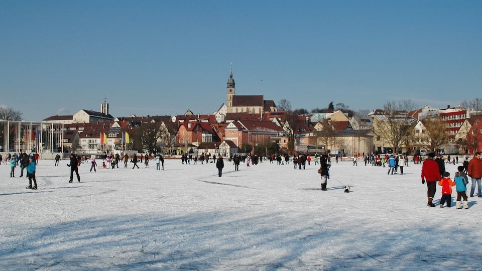A snowy square with people skating and walking, surrounded by historic buildings and a church tower.