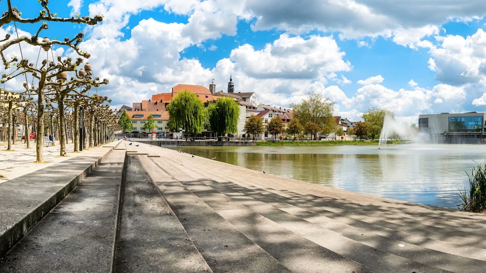 A riverside walkway with trees, a fountain, and buildings in the background.