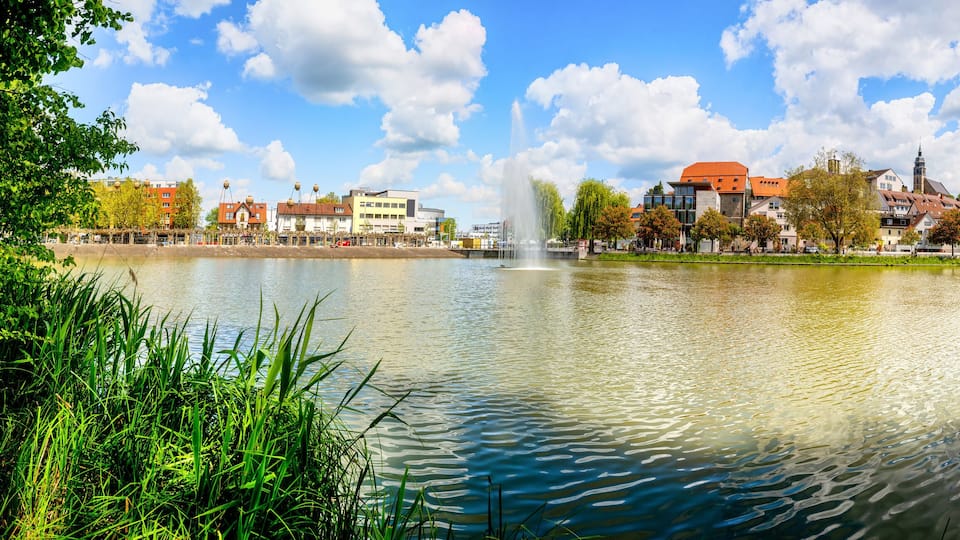 A lake with a fountain, surrounded by trees and buildings.