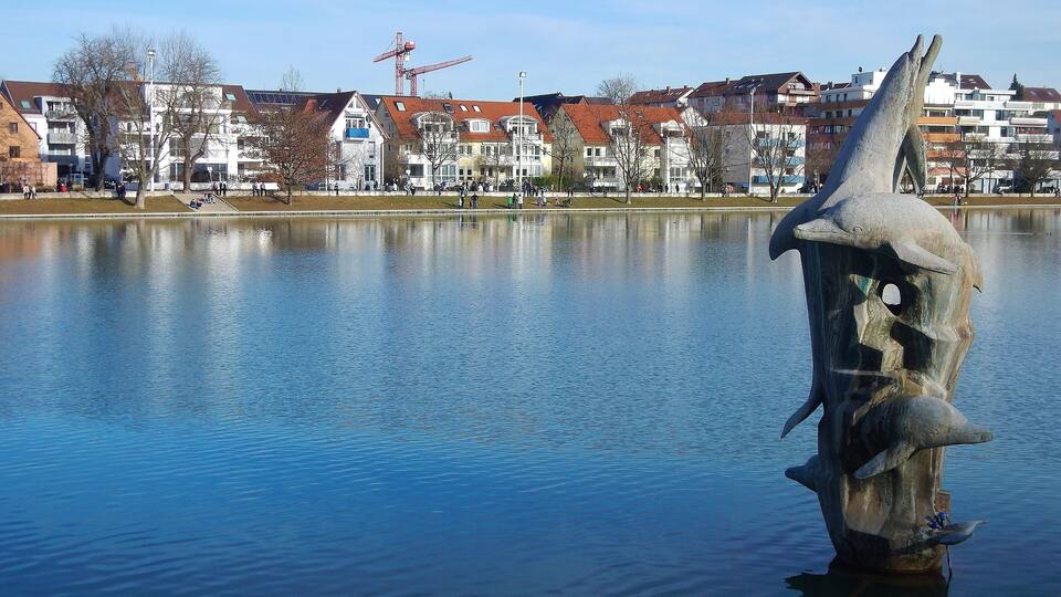 A calm lake with a sculpture of a fish in the foreground and residential buildings in the background.