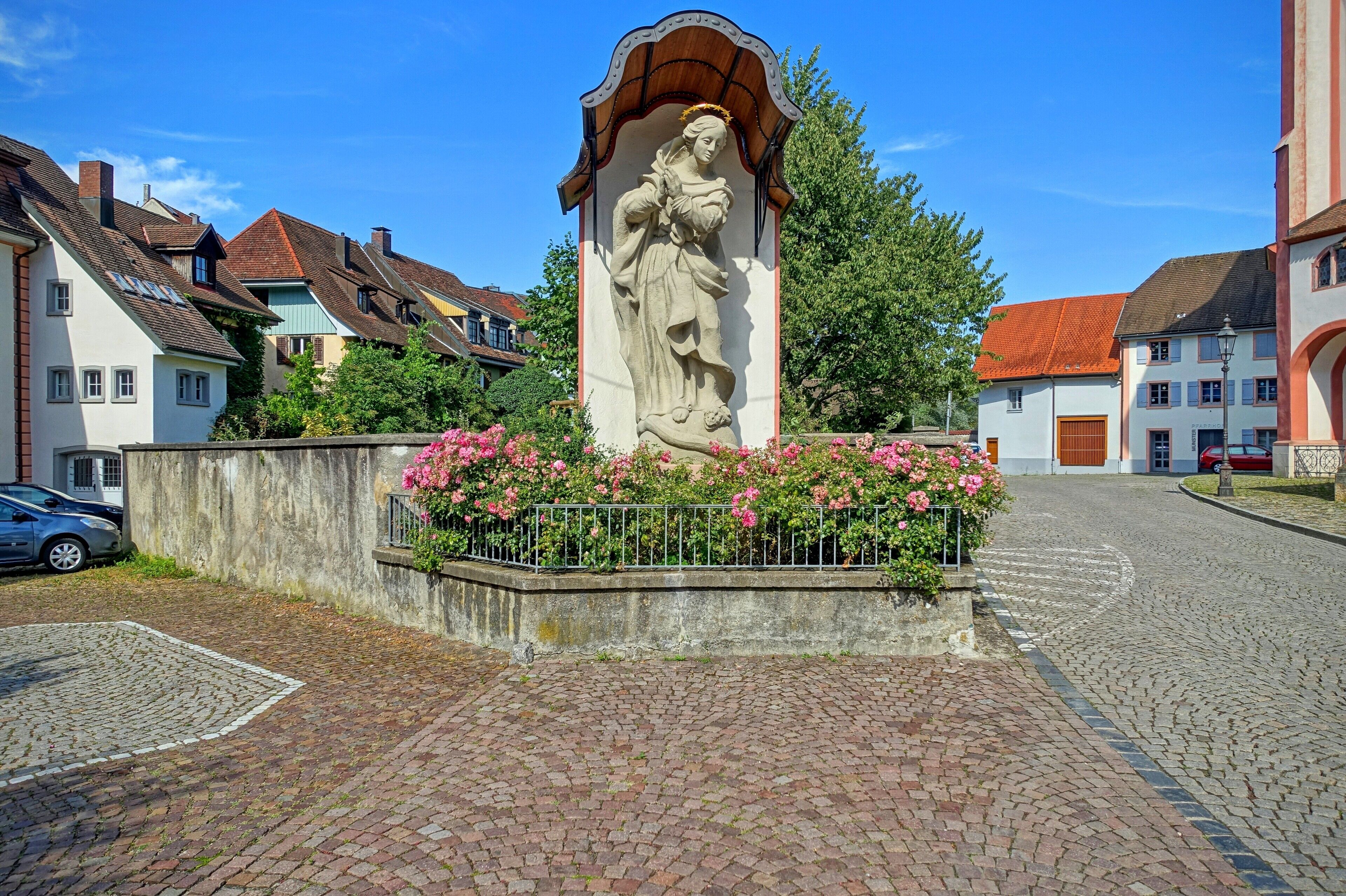 Die Marienstatue steht an der Nordseite des Münsters vor der Mauer des Pfarrgartens. Früher befand sich hier der Friedhof. Die Statue wird durch eine kunstvoll geschwungene Überdachung und eine Rückwand geschützt. Die etwa vier Meter hohe Marienstatue wurde 1727 geschaffen und in der Fassadenmitte zwischen den Münstertürmen aufgestellt. Sie hat den Brand des Münsters im Jahr 1751 überstanden. Starke Verwitterung hatte den Sandstein dieser Statue so stark geschädigt, dass sie durch eine Kopie ersetzt werden musste. Am jetzigen Standort steht die Madonna von 1727 seit dem Jahr 1913. Durch mehrere fachgerechte Restaurierungen ist nicht mehr erkennbar, dass es sich hier um die Original-Statue handelt. Kunstgeschichtlich wird dieser Madonnentypus als "Immaculata" (Der unbefleckten Empfängnis) bezeichnet. Das Haupt Marias ist von zwölf Sternen umkränzt, die Schlange als Symbol der Sünde, die sich um die Weltkugel windet, wird von Maria zertreten. Die Zahl zwölf steht für: die zwölf Stämme Israels, die zwölf Apostel, die zwölf Tierkreiszeichen, die zwölf Monate – symbolhaft für Vollkommenheit und Vollständigkeit. Da die Europafahne am 8. Dezember 1955, einem Marienfeiertag, vom Ministerrat in der bestehenden Form bestimmt wurde, wird sie in Verbindung mit Maria gebracht. Besonders die blaue Farbe und auch die zwölf Sterne sind ein symbolhafter Hinweis. Die zwölf goldenen Sterne auf blauem Hintergrund versinnbildlichen sämtliche europäischen Nationen, eben als Symbol der Vollständigkeit. Die Marienstatue hat der aus Bayern stammende Bildhauer Michael Speer im Jahr 1727 geschaffen. Der graue Sandstein stammt aus dem Fricktal.
