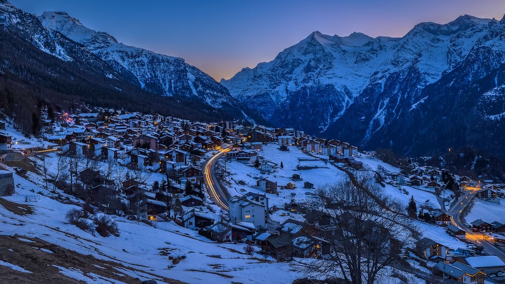 A snow-covered mountain village at dusk with illuminated buildings and winding roads.