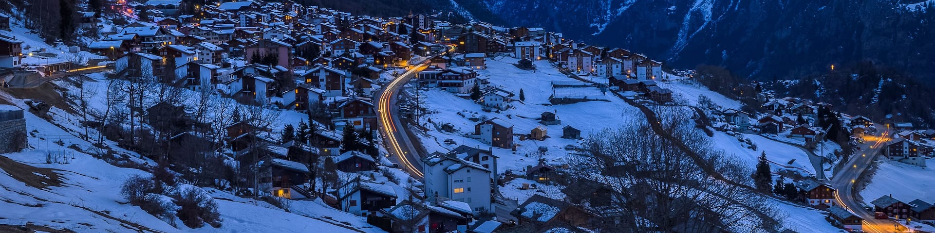 A snow-covered mountain village at dusk with illuminated buildings and winding roads.