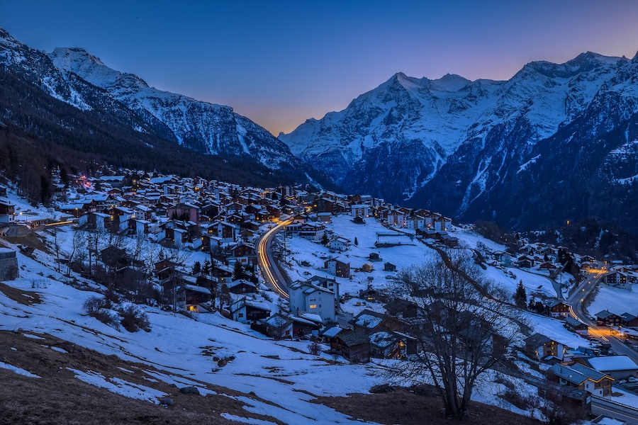 Un village de montagne enneigé, baigné par la lumière du crépuscule, avec des bâtiments illuminés et des routes sinueuses.