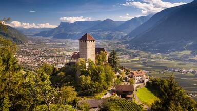 A historic tower on a hill overlooking a valley with vineyards, surrounded by mountains.