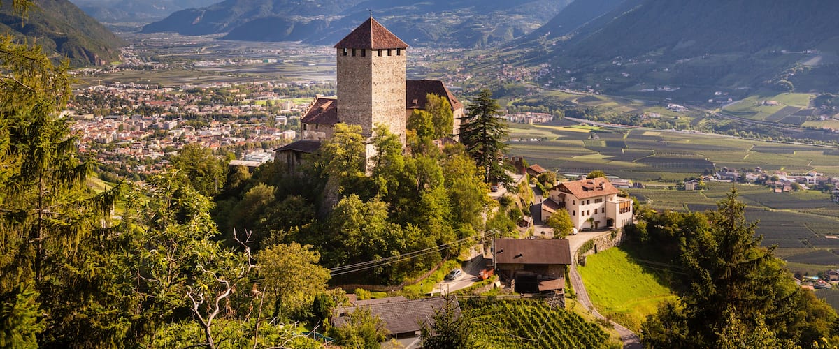 A historic tower on a hill overlooking a valley with vineyards, surrounded by mountains.