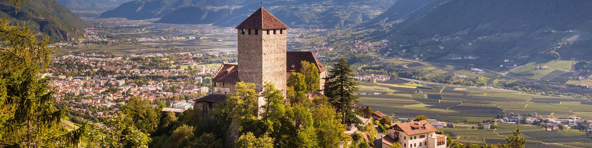 A historic tower on a hill overlooking a valley with vineyards, surrounded by mountains.