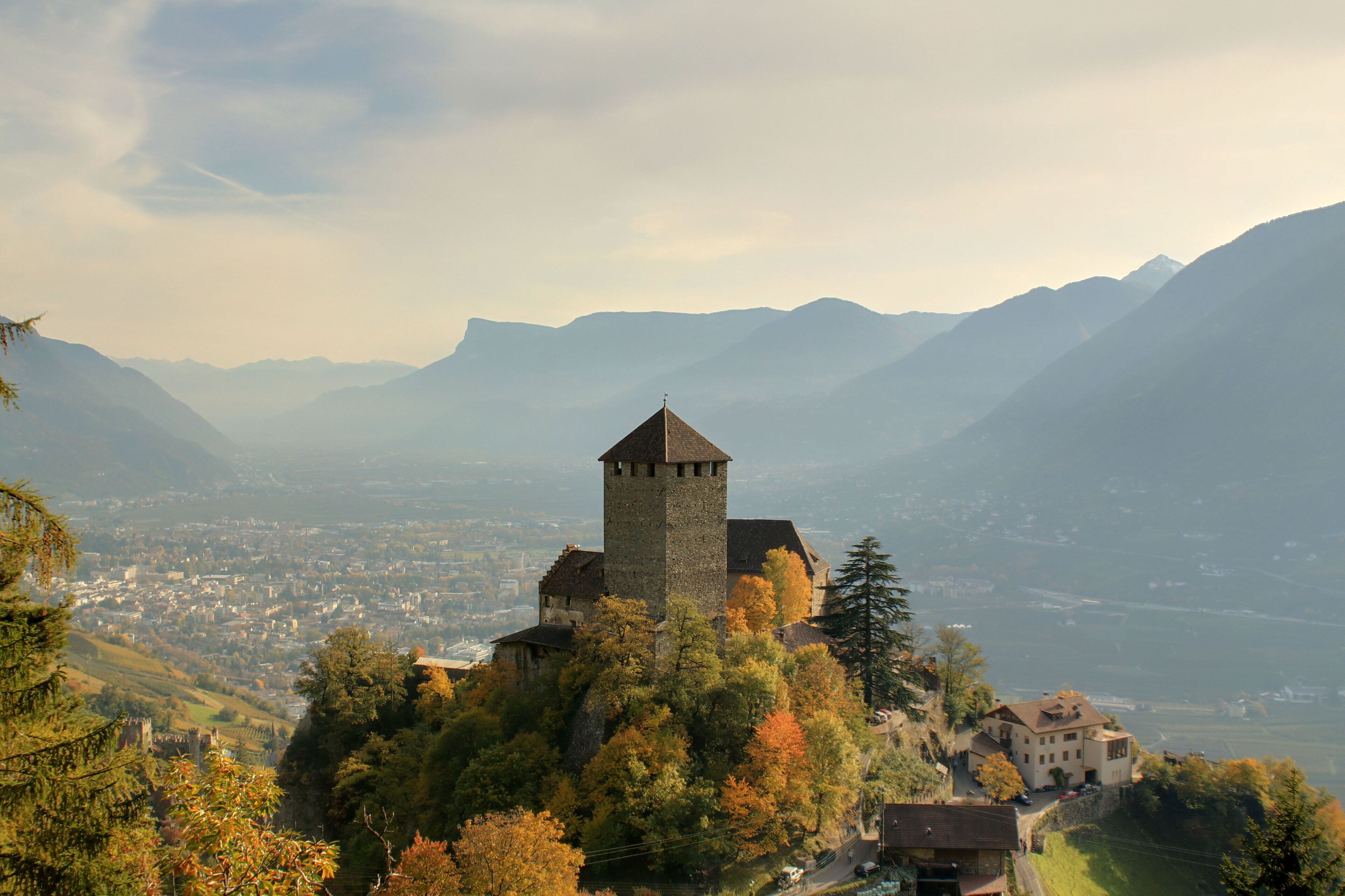 A castle on a hill with autumn foliage, surrounded by mountains and a valley below.