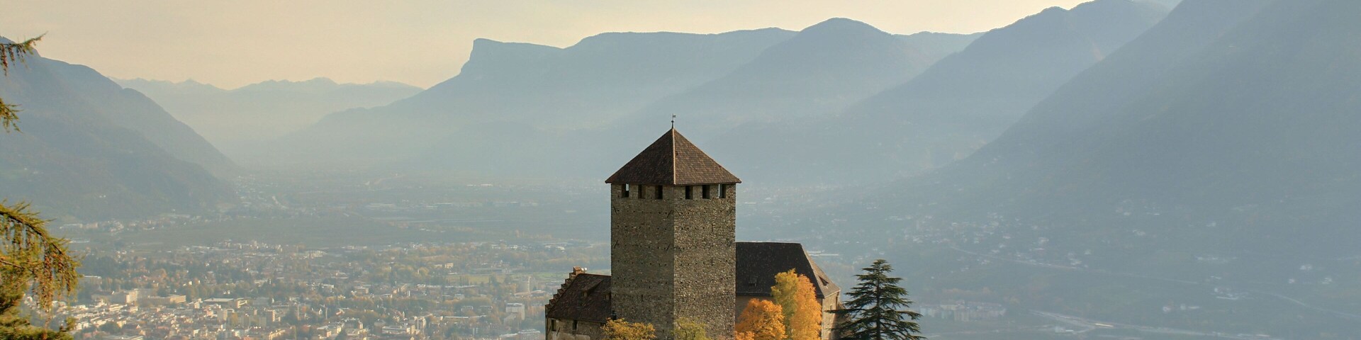 Un castillo en una colina rodeado de follaje otoñal, montañas y un valle en la parte inferior.