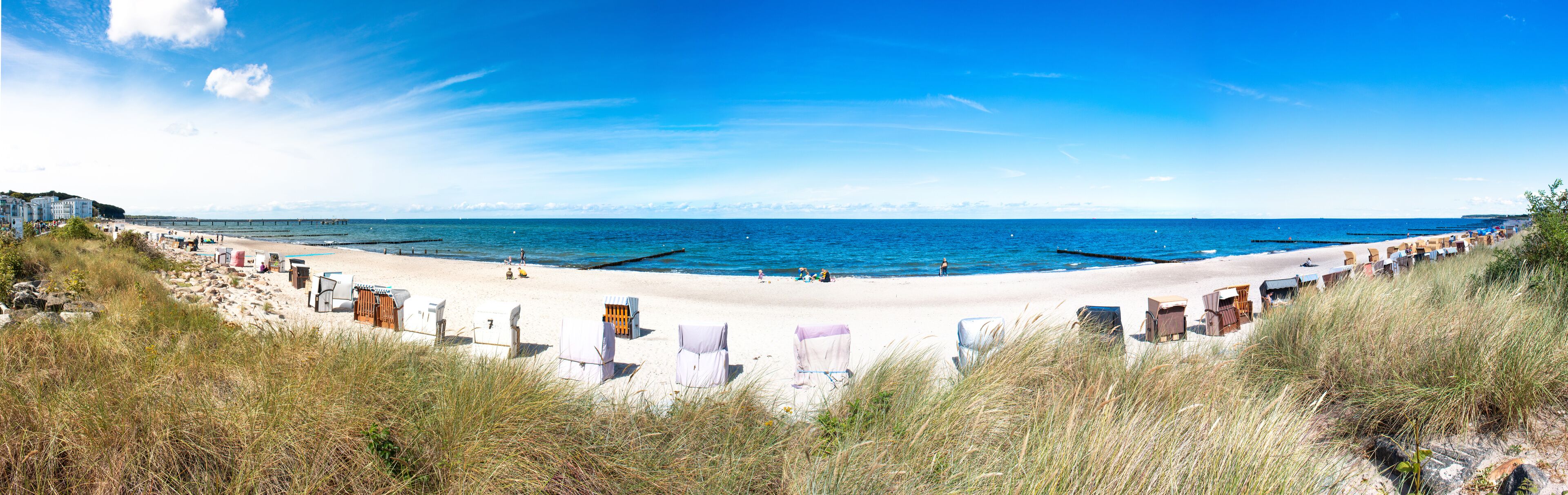 A beach with white sand, a pier in the distance, and grassy dunes in the foreground.