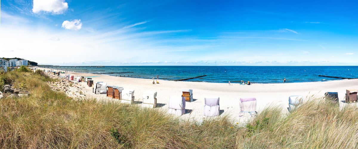 A beach with white sand, a pier in the distance, and grassy dunes in the foreground.