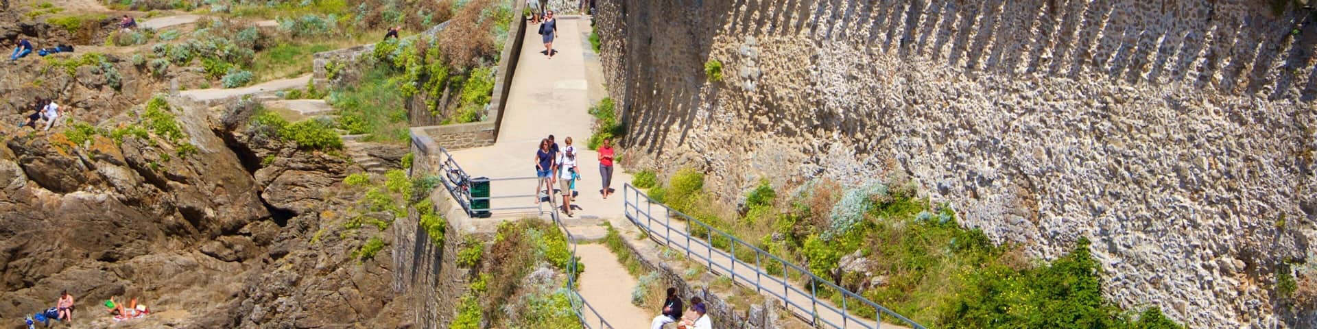 A historic stone fortification with a sea view, a paved pathway, and a modern building with balconies.