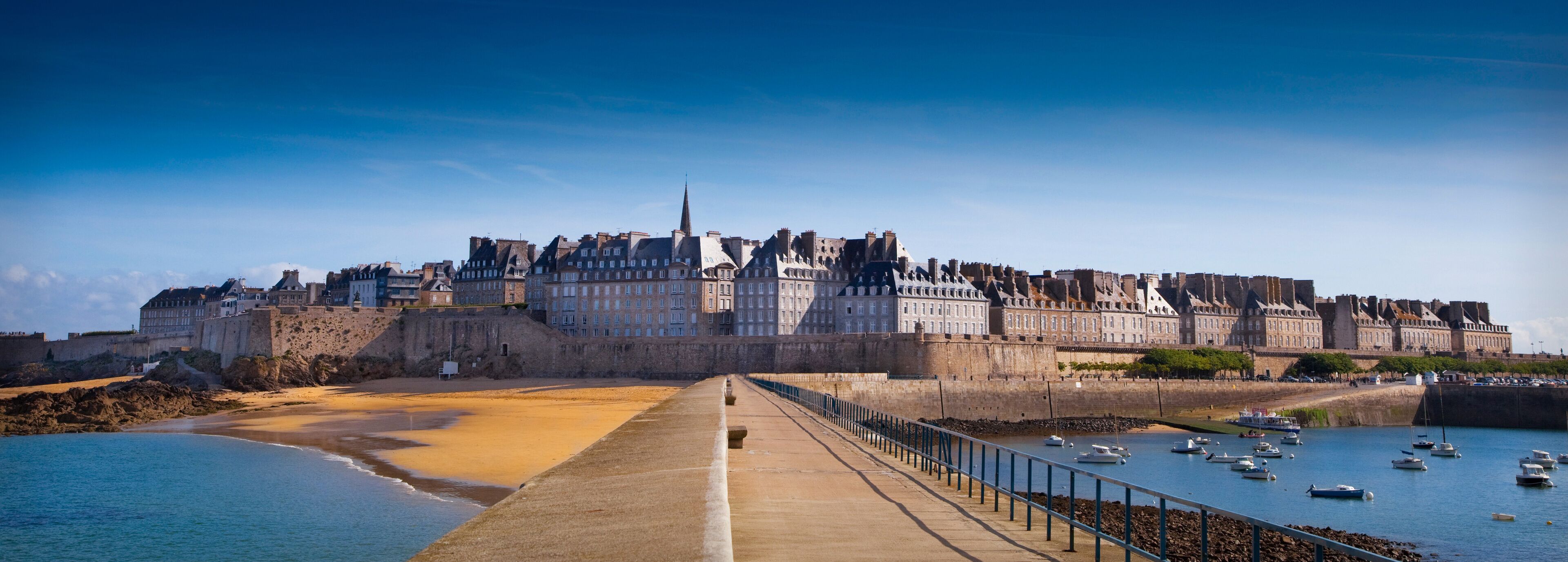 A coastal city with a prominent castle, sandy beach, and a promenade lined with boats.