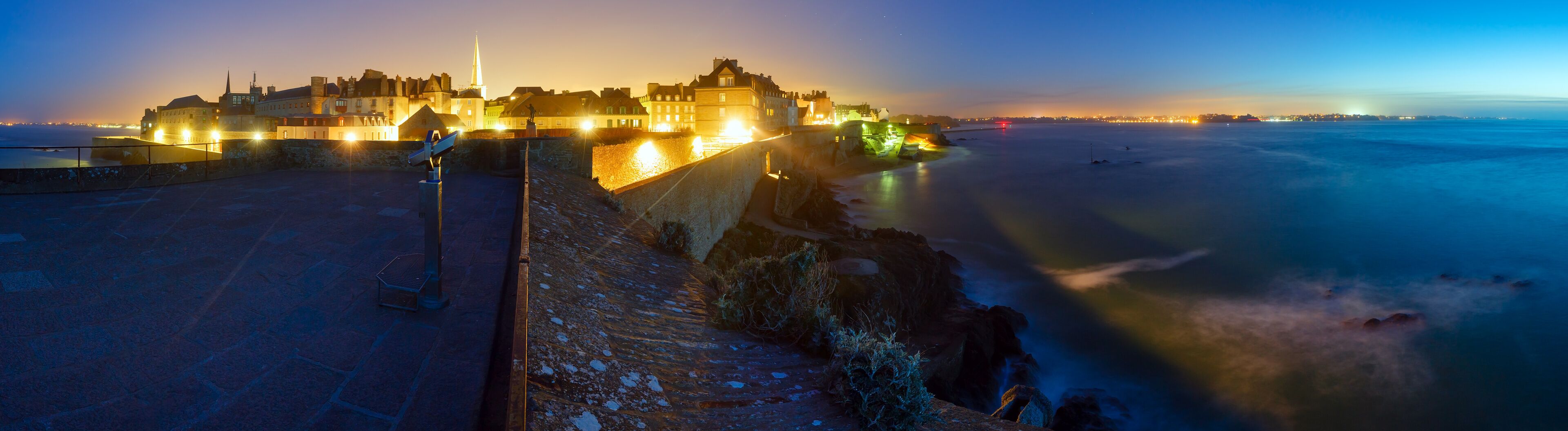 A coastal cityscape at dusk with illuminated buildings and a prominent castle.