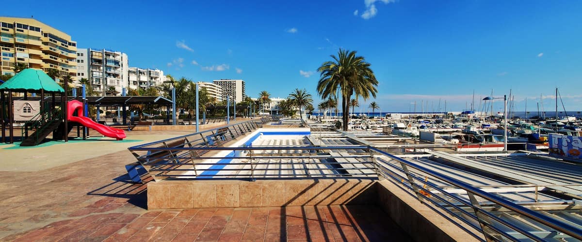 A marina with boats, a playground with a red slide, palm trees, and buildings in the background.