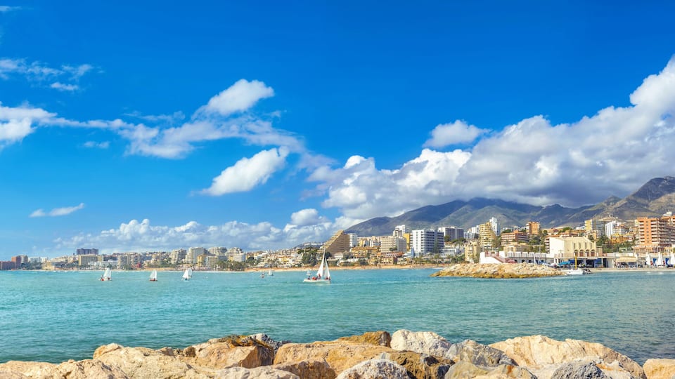 A coastal city with sailboats on the water, rocky shoreline, and a mountain range in the background.