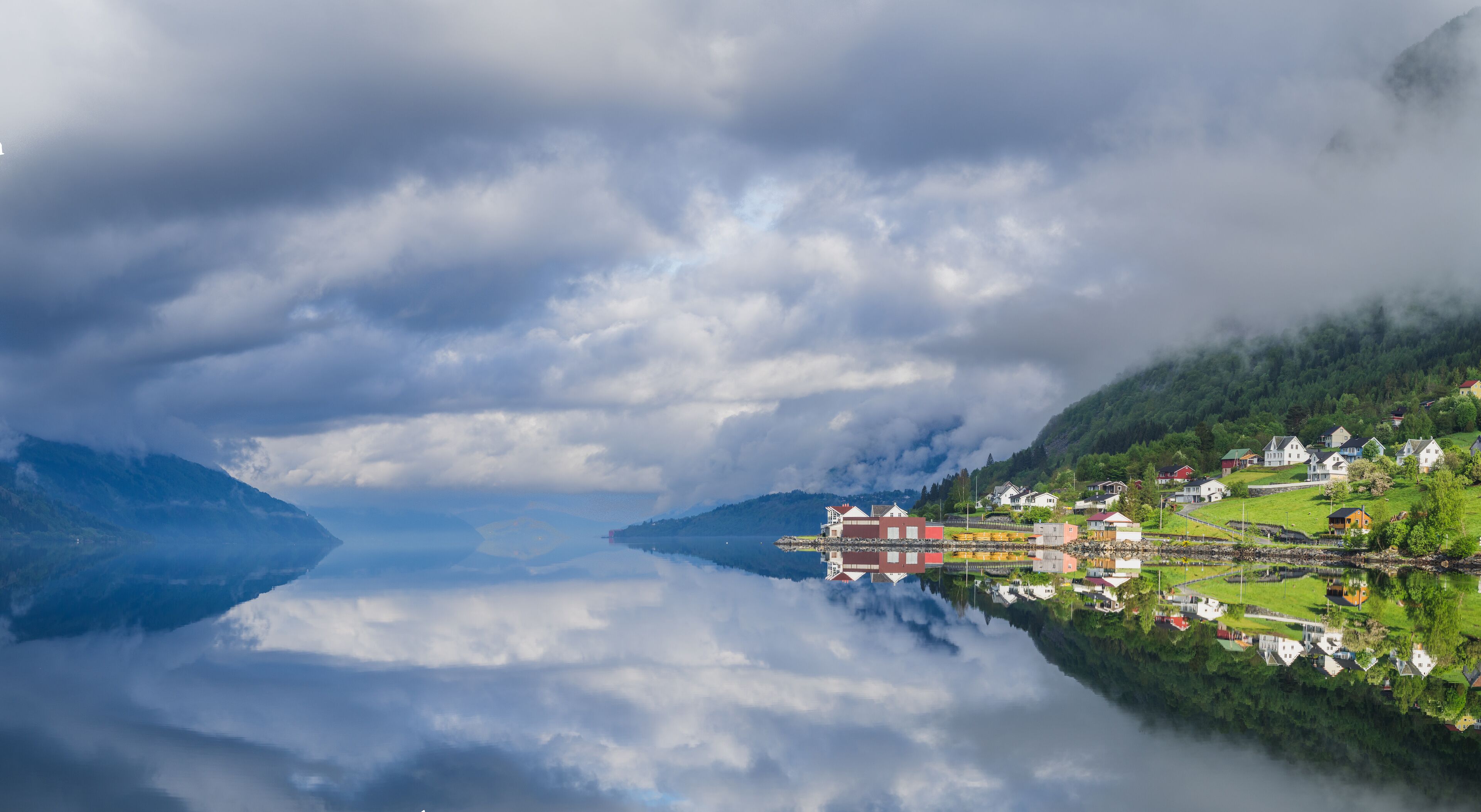 A serene lakeside village with houses reflecting on calm water, surrounded by lush greenery and mountains.