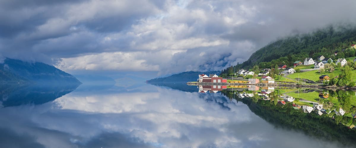 A serene lakeside village with houses reflecting on calm water, surrounded by lush greenery and mountains.