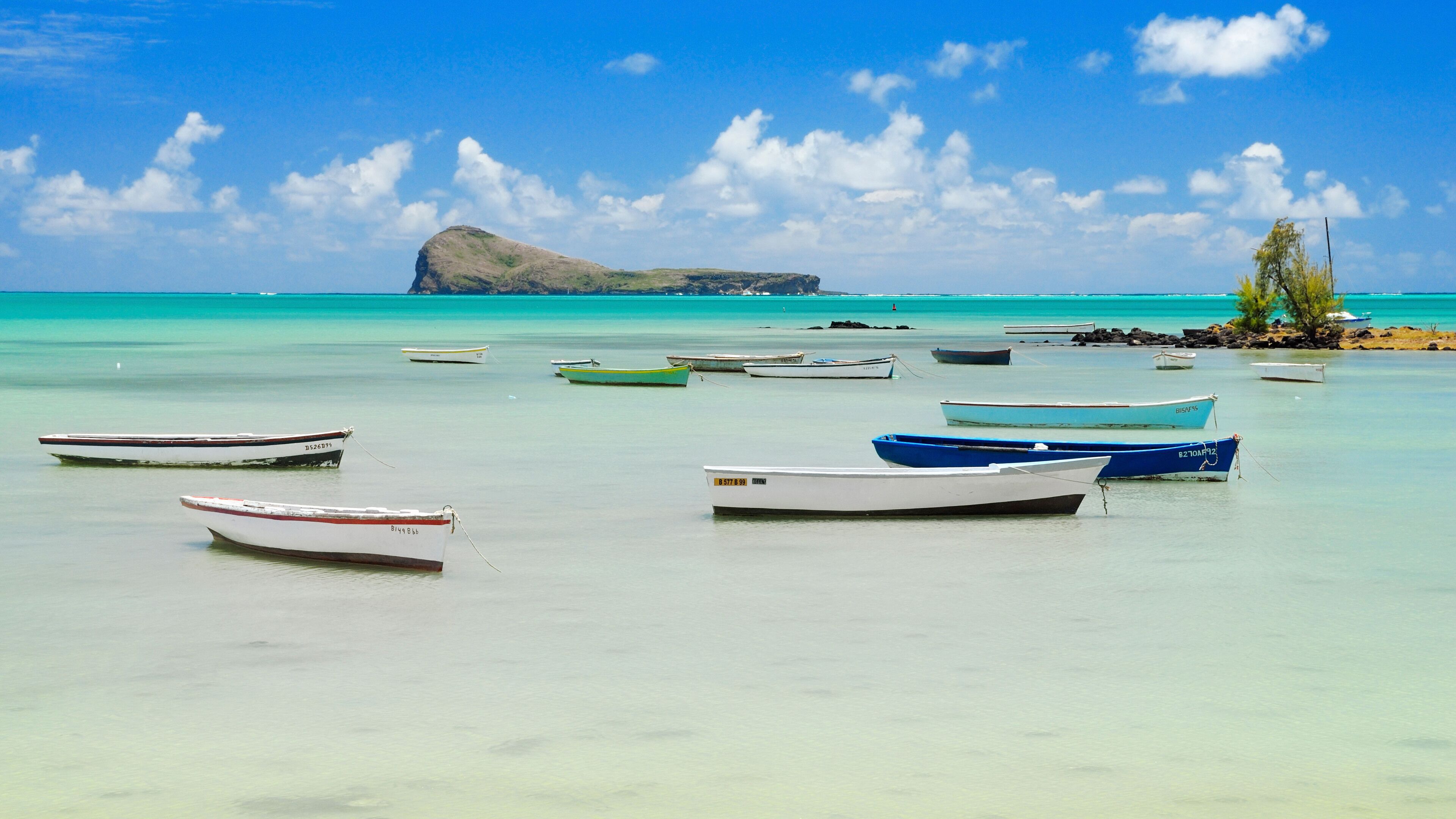 A turquoise sea with several small boats anchored near the shore, a rocky outcrop in the background, and a clear blue sky.