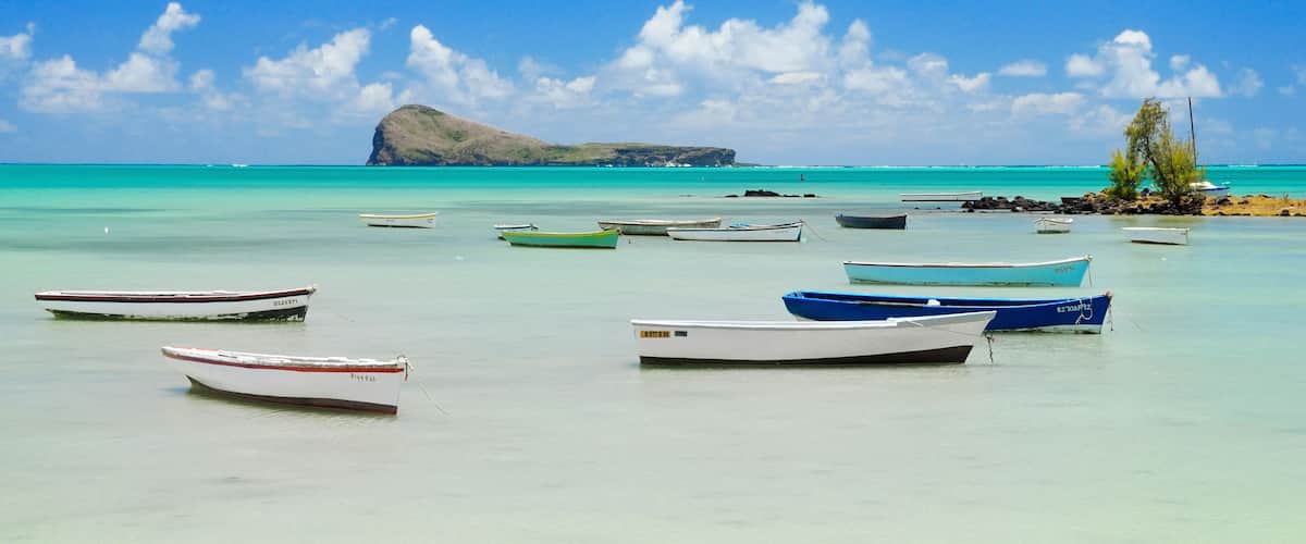 A turquoise sea with several small boats anchored near the shore, a rocky outcrop in the background, and a clear blue sky.