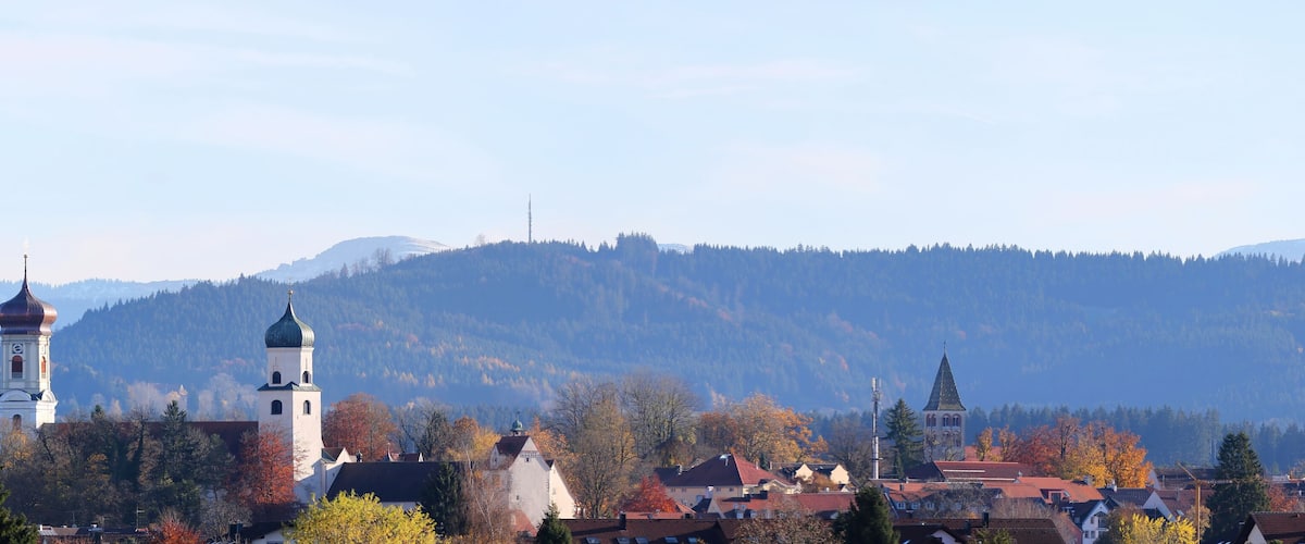 A town with a prominent church tower, surrounded by autumn-colored trees and mountains in the background.