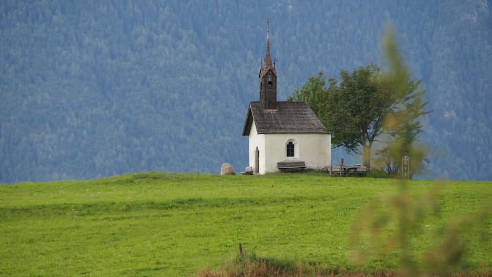 A small white church with a steeple on a grassy hill with a forested mountain in the background.